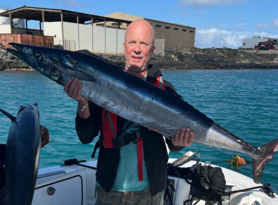 In der Region São Vicente, Kapverden, präsentiert ein Angler stolz seinen großen Fang eines Wahoos auf einem Boot, umgeben von klarem Wasser. Perfekter Angelurlaub für Meeresangeln-Liebhaber.