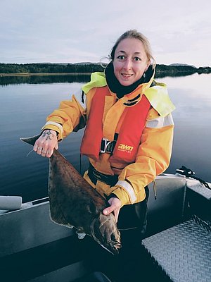 Eine Anglerin hält einen gefangenen Fisch an Bord eines Bootes vor der malerischen Kulisse von Hitra, Norwegen. Perfekte Bedingungen für einen erfolgreichen Angelausflug im ruhigen Gewässer. Angelurlaub, Naturerlebnis, Meeresangeln.