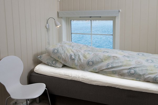 Ein schlicht eingerichtetes Zimmer mit einem Bett und einem Stuhl hat ein Fenster mit Blick auf das ruhige, blaue Wasser.