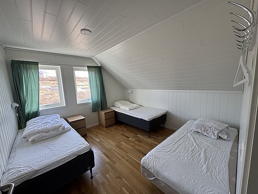 Gemütliches Schlafzimmer in Hitra, Norwegen: Drei Betten, Holzfußboden, weiße Wandverkleidung, zwei Fenster mit grünen Vorhängen, ideal für komfortablen Angelurlaub. Entspannte Atmosphäre in skandinavischem Stil. Perfekt für Erholung nach erlebnisreichen Angeltagen.