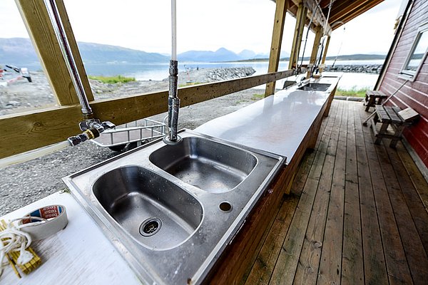 Außenküche in Nord-Norwegen: Holzveranda mit Edelstahlspülen, Blick auf Fjord und Berge. Perfekt für Angler, um den Fang frisch zuzubereiten. Ideal für Angelurlaub und Naturerlebnis in atemberaubender Umgebung.