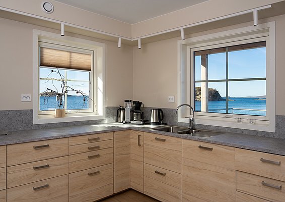 Moderne Küche mit Holzschränken und Meerblick in Hitra, Norwegen. Helle Fenster lassen das Tageslicht herein, ideal für entspannte Momente nach einem erfolgreichen Angelausflug. Perfekte Kombination aus Komfort und Naturerlebnis.