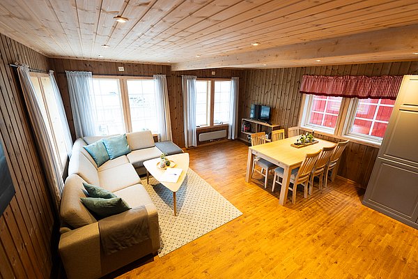Gemütliches Wohnzimmer in Nord-Norwegen mit Holzwänden, heller Sofaecke, Esstisch für sechs Personen. Ideal für Angelurlaub. Große Fenster bieten viel Tageslicht und Aussicht auf die Umgebung. Perfekte Unterkunft für Erholung nach aufregenden Angeltagen.