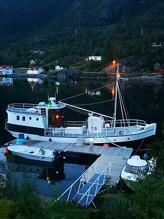 Abendstimmung in Nord-Norwegen: Historisches Boot am ruhigen Fjordhafen, umgeben von malerischen, bewaldeten Hängen. Perfekter Ausgangspunkt für Angelreisen und Meeresangeln inmitten beeindruckender Natur.
