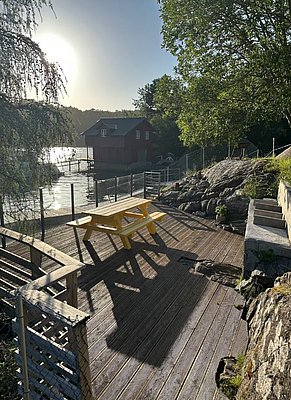 Holzterrasse in Hitra, Norwegen: Ein idyllischer Blick auf ein ruhiges Gewässer, umgeben von einem sonnigen Pier, Bäumen und einem charmanten roten Bootshaus – perfekt für entspannte Angel- und Naturerlebnisse.