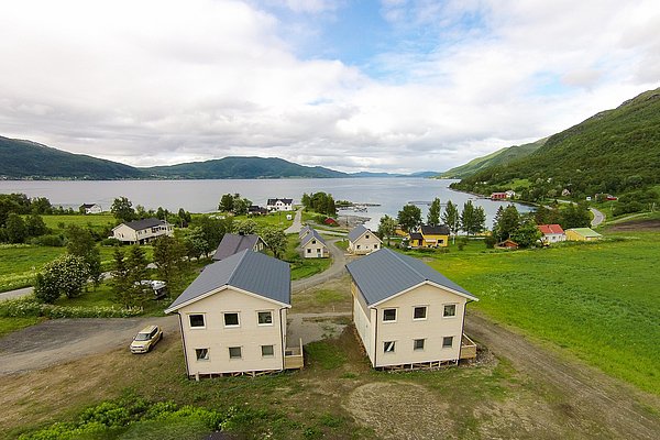 Luftaufnahme einer idyllischen Landschaft mit mehreren nebeneinander stehenden Häusern, umgeben von grünen Feldern, hügeligen Bergen und einem ruhigen See im Hintergrund. Luftaufnahme einer idyllischen Landschaft mit mehreren nebeneinander stehenden Häusern, umgeben von grünen Feldern, hügeligen Bergen und einem ruhigen See im Hintergrund.