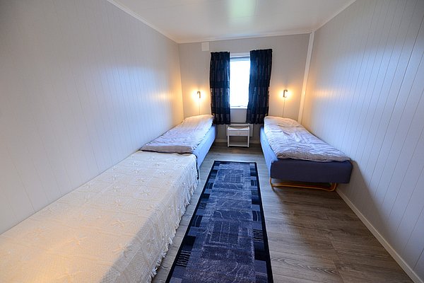 Gemütliches Zimmer mit zwei Einzelbetten in Nord-Norwegen. Helle Wände, Holzfußboden und ein Fenster mit Vorhängen schaffen eine einladende Atmosphäre. Ideal für Erholung nach einem erfolgreichen Angeltag in atemberaubender Natur.