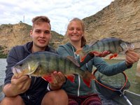 Zwei Personen sitzen in einem Boot und halten stolz Flussbarsche vor einer felsigen Kulisse in die Kamera.
