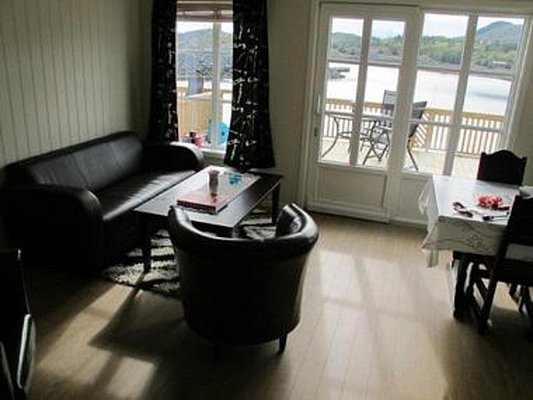 Gemütliches Wohnzimmer in Nordland, Norwegen, mit Ledersofas, Tisch und Stühlen vor großen Fenstern. Heller Holzboden mit Terrasse und Blick auf Fjordlandschaft. Perfekt für Entspannung nach einem erfolgreichen Angeltag.
