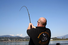 Ein Angler in schwarzem Pullover, mit einer stark gebogenen Angelrute, fischt an einem See vor einer malerischen Berglandschaft.