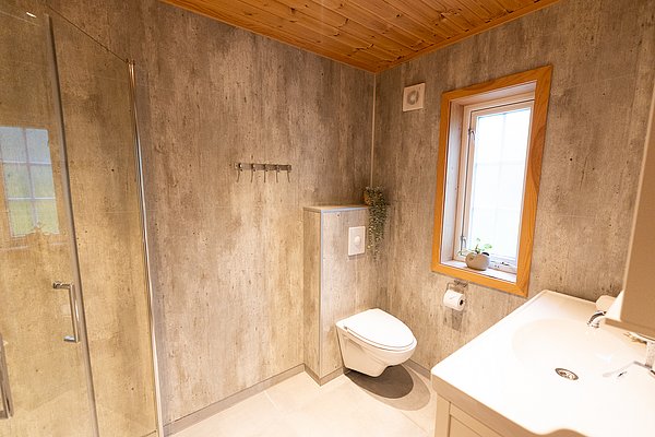 Modernes Badezimmer in Nord-Norwegen: Helle Fliesen, hängendes WC, Duschkabine, Fenster mit Holzeinfassung, Pflanzen sorgen für Wohlfühlatmosphäre. Ideal für erholsame Angelreisen.