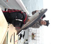 Eine Person in Winterkleidung hält auf einem Boot einen großen, länglichen Fisch vor einer Meereskulisse.