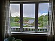 Blick aus einem Fenster auf die malerische Landschaft Nordlands, Norwegen. Zu sehen sind ein ruhiger Fjord, umgeben von grünen Hügeln und roten Holzhäusern. Perfekte Kulisse für einen entspannenden Angelurlaub.