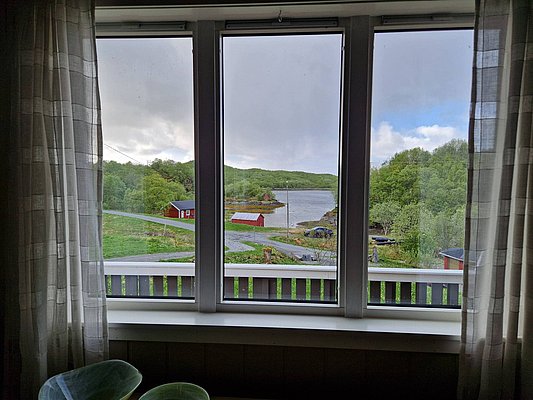 Blick aus einem Fenster auf die malerische Landschaft Nordlands, Norwegen. Zu sehen sind ein ruhiger Fjord, umgeben von grünen Hügeln und roten Holzhäusern. Perfekte Kulisse für einen entspannenden Angelurlaub.