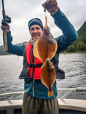 Ein Angler mit rot-blauer Jacke hält stolz zwei frisch gefangene Plattfische auf einem Boot, vor bewaldeter Küste in Norwegen. Perfekter Angelurlaub in Hitra für Abenteuer und reiche Fischgründe.