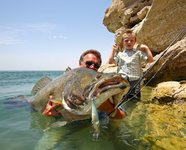 Ein Mann und ein Junge stehen im flachen Wasser an einem felsigen Ufer und halten einen großen Fisch, vermutlich einen Bass, in die Kamera.