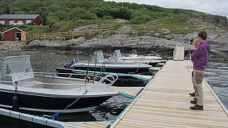 Angelboote an einem Holzsteg in der malerischen Landschaft Nord-Norwegens. Idealer Ausgangspunkt für Meeresangeln-Abenteuer. Eine Person schaut auf die Boote, umgeben von grünen Hügeln und Küstenlandschaft.