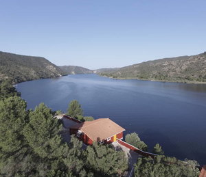 Luftaufnahme des Riba-Roja-Stausees in Spanien: glitzerndes Wasser, umgeben von grünen Hügeln und dichter Vegetation. Ein rotes Gebäude mit Ziegeldach am Ufer, idealer Ausgangspunkt für Angelabenteuer in atemberaubender Natur.