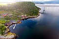 Luftaufnahme von Nordland, Norwegen: Malerische Küstenlandschaft mit grünen Hügeln, roten Häusern, einer Brücke und einem Steg am ruhigen Wasser. Ideal für Angelreisen und Naturerlebnisse.