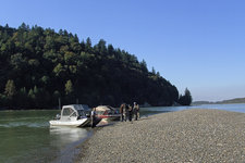 Menschen stehen neben Booten auf einer Kiesbank am Fluss, umgeben von bewaldeten Hügeln und klarem Himmel.