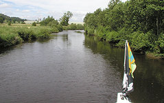 Ein ruhiger Fluss mit bewachsenen Ufern und einer Bootsspitze mit Flaggen am rechten Bildrand.