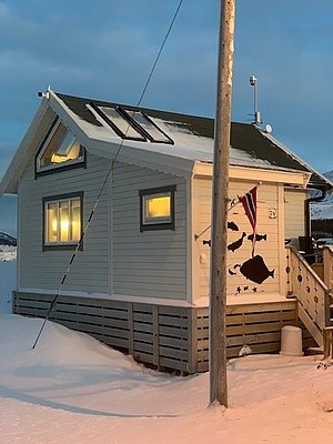 Kleines, gemütliches Holzhaus in verschneiter Landschaft Nord-Norwegens. Eine norwegische Flagge und Fischdekoration zieren die Außenwand. Ideal für Angelurlaub und Erholung, umgeben von winterlicher Ruhe und skandinavischer Gemütlichkeit. Perfekter Rückzugsort im hohen Norden.