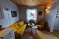 Gemütliches Wohnzimmer in Hitra, Norwegen: Gelbes Sofa, gelbe Sessel und Holztisch. Weiß getäfelte Wände, Holzboden, Fenster mit Ausblick in den Garten. Perfekter Ort zum Entspannen nach einem Tag Angeln.