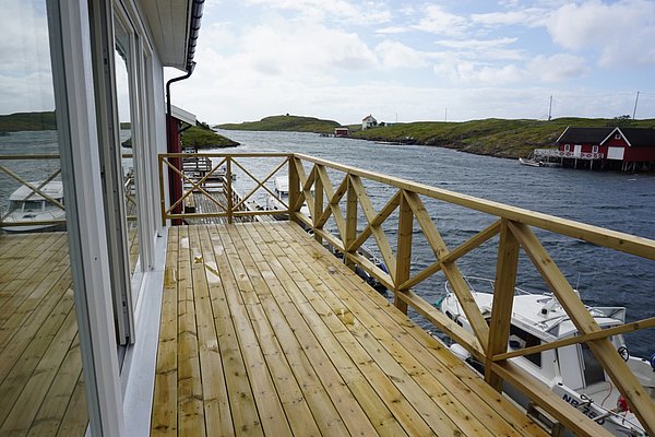 Große Holzterrasse mit Blick auf norwegische Küstenlandschaft und ruhige Gewässer. Rote Fischerhütten und Boote im Wasser. Perfekt für Angelurlaube und Naturerlebnisse in Nordland, Norwegen. Ideal für Entspannung und Abenteuer zugleich.