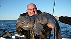 Ein Angler hält stolz einen großen, gefangenen Fisch an Bord eines Boots in Nord-Norwegen. Der klare Himmel und das blaue Meer im Hintergrund betonen das Abenteuer Meeresangeln.