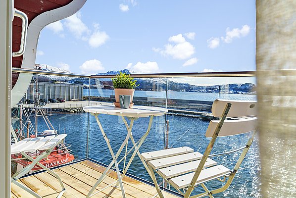 Ein sonniger Balkon mit weißem Tisch und Stühlen, Blick auf den Fjord und schneebedeckte Berge in Südwest-Norwegen. Ideal für Angelurlaub und Entspannung.
