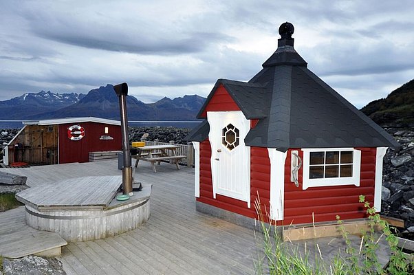 Rote Holzhütte mit traditioneller Architektur in Nord-Norwegen, umgeben von malerischen Bergen am Meer. Perfekter Ausgangspunkt für Angelurlaub und Naturerlebnisse im hohen Norden. Stimmungsvoll mit rauer, unberührter Landschaft im Hintergrund.