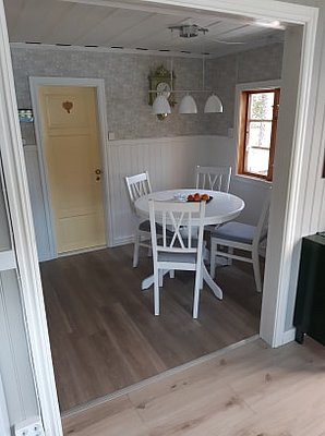 Ein gemütliches Esszimmer mit einem runden weißen Tisch, vier weißen Stühlen und einer Schale mit Früchten auf dem Tisch, umgeben von hellen Wänden und Holzboden.