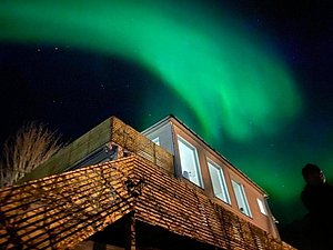 Ein modernes Holzhaus in Nord-Norwegen wird bei Nacht von beeindruckenden, schimmernden Nordlichtern erleuchtet. Eine klare Sternennacht verstärkt die magische Atmosphäre im hohen Norden.