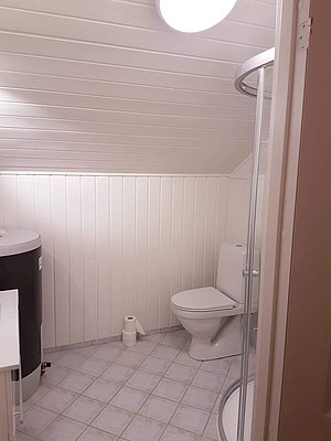 Helles, modernes Badezimmer in Nord-Norwegen mit Dusche, Toilette und gefliestem Boden. Die weißen Holzpaneele verleihen dem Raum eine gemütliche Atmosphäre und laden zum Entspannen nach einem ereignisreichen Angelabenteuer ein.