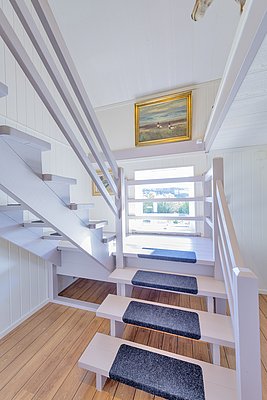 Helles Treppenhaus in Nordland, Norwegen mit Holzböden und weiß gestrichenen Wänden. Ein Bild in Goldrahmen dekoriert die Wand, während ein Fenster idyllische Ausblicke bietet. Skandinavische Innenarchitektur trifft auf natürliche Schönheit.