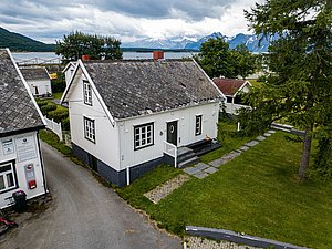 Weiße Holzhäuser in Nord-Norwegen, malerisch eingebettet in grüner Landschaft, mit spektakulärem Blick auf ein blaues Fjord und majestätische Berge im Hintergrund. Perfekter Ausgangspunkt für Angelreisen und Erkundungstouren.