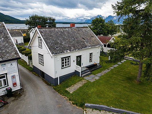 Weiße Holzhäuser in Nord-Norwegen, malerisch eingebettet in grüner Landschaft, mit spektakulärem Blick auf ein blaues Fjord und majestätische Berge im Hintergrund. Perfekter Ausgangspunkt für Angelreisen und Erkundungstouren.