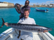 Zwei Angler präsentieren stolz ihre Fänge auf einem Boot vor der Küste von São Vicente, Kapverden. Der türkisfarbene Ozean und ein Fischerboot im Hintergrund schaffen eine perfekte Atmosphäre für Angelabenteuer.