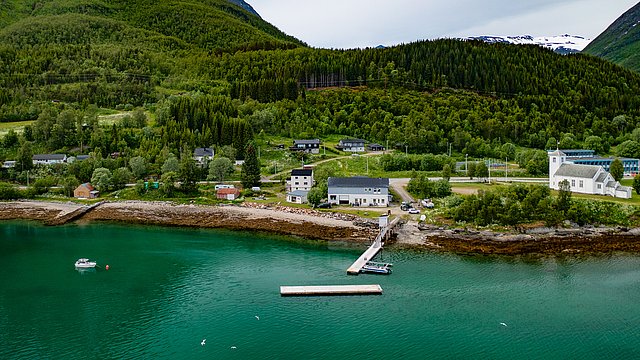 Luftaufnahme eines idyllischen Küstendorfs in Nord-Norwegen, mit grünen Hügeln, einem Steg und kristallklarem Wasser. Perfekt für Angelreisen und Naturerlebnisse in malerischer Umgebung. Entspannung und Abenteuer erwarten Reisende hier.