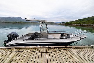Ein modernes Angelboot liegt an einem Holzsteg in Nord-Norwegen. Im Hintergrund sind ruhige, klare Gewässer und bewaldete Hügel unter bewölktem Himmel zu sehen. Perfekter Ausgangspunkt für Angelabenteuer.