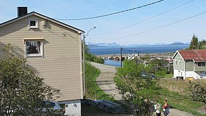 Idyllische Küstenszene in Nord-Norwegen: malerische Dorfhäuser mit Blick auf das Meer, umgeben von grüner Landschaft und majestätischen Bergen am Horizont. Perfekte Kulisse für Angelreisen und Erkundungstouren.