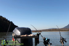 Das Bild zeigt Angelruten und einen Motor auf einem Boot in einem malerischen Gewässer umgeben von bewaldeten Hügeln und klarem Himmel.