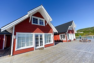 Zwei rote Ferienhäuser im traditionellen Stil stehen auf einer Terrasse mit Blick auf das klare Wasser und grüne Hügel in Nord-Norwegen. Perfekt für Angelreisen und naturnahe Erholung geeignet, unter strahlend blauem Himmel.