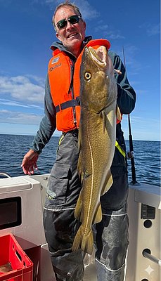 Ein Angler hält stolz einen großen Kabeljau an Bord eines Bootes vor der beeindruckenden Kulisse der Norwegischen See bei Hitra. Perfektes Angelabenteuer bei strahlendem Sonnenschein und klarem Himmel.