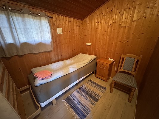 Gemütliches Schlafzimmer in Holzhütte auf Hitra, Norwegen. Enthält ein Einzelbett, Nachttisch, Lampe, Teppich und Fenster mit Vorhang. Ideal für Angelabenteuer in natürlicher Umgebung.