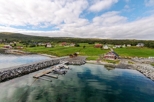Luftaufnahme eines norwegischen Küstendorfs in Nord-Norwegen, mit malerischen bunten Häusern, grünen Feldern und einem ruhigen Bootsanleger am klaren Fjordwasser. Perfekt für Angelurlaube und Naturerlebnisse.