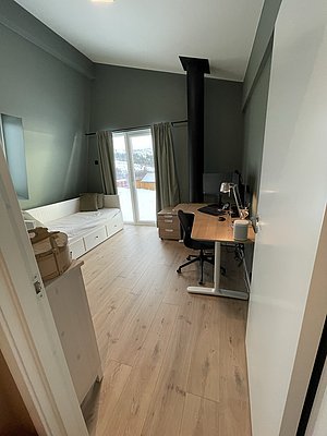 Gemütliches Zimmer in Hitra, Norwegen: Holzboden, Schreibtisch, Tagesbett, Ofen. Blick auf verschneite Landschaft durch große Fenster. Ideal zum Entspannen nach einem erlebnisreichen Angeltag.