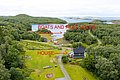 Luftaufnahme der Region Nordland, Norwegen: Grüne Landschaft mit einem Haus, dahinter Boote und ein Filetierhaus nahe der Küste. Ideal für Angelurlaub und Outdoor-Abenteuer in malerischer Umgebung.