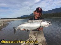 Ein Angler hält stolz einen großen Fisch am Flussufer, umgeben von schöner Berglandschaft.