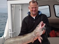Ein Mann hält lächelnd einen großen Seehecht auf einem Boot vor einer Meereskulisse.